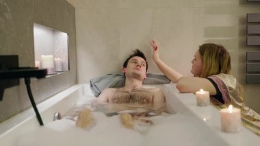 Genç bir adam köpük banyosunda kafasını havluya yaslayıp dinlenirken, bir kadın da mum ışığında saçlarını nazikçe okşuyor. Yüksek kalite 4k görüntü