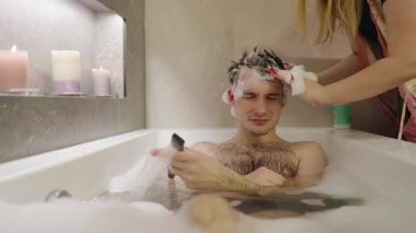 Genç bir adam köpük banyosunda rahatlarken kız arkadaşı saçına şampuan sürüp rahatlatıcı ve samimi bir an yaratıyor. Yüksek kalite 4k görüntü