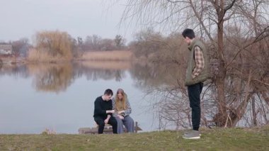 Üç genç insan gölün kenarında, bir adam ayakta dururken bir oğlan ve bir kız, düşüncelere dalmış, ahşap bir rıhtımda oturuyor. Yüksek kalite 4k görüntü