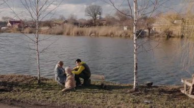 Yaşlı bir kadın ve torunu, sıcak bir bahar gününde nehir kenarında Golden Retriever 'larını okşayarak oturuyorlar. Sakin su arka plandaki köy ve kiliseyi yansıtır. Yüksek kalite 4k görüntü