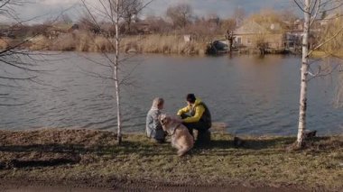 Son sınıf öğrencisi bir kadın ve torunu nehrin kenarında Golden Retriever 'larını okşayarak huzurlu bir anın tadını çıkarıyorlar. Kırsal bölgede parlak bir gökyüzünün altında bir köy ve uzak bir kilise bulunur. Yüksek kalite 4k