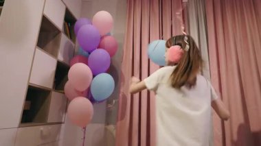 Genç bir kız, pembe perdeleri ve kulaklıkları olan neşeli bir iç mekan ortamında balonlarla mutlu bir şekilde oynuyor. Yüksek kalite 4k görüntü