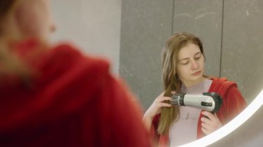 Kırmızı bornozlu genç bir kadın aynadaki yansımasına bakarken saçını saç kurutma makinesiyle kurutuyor. Yüksek kalite 4k görüntü