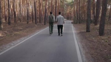 İki adam bir Golden Retriever 'la çam ormanında asfalt bir yolda yürüyor. Yan yana yürürler, doğanın huzurlu atmosferinin ve önlerindeki açık yolun tadını çıkarırlar. Yüksek kalite 4k görüntü