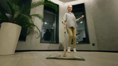 Orta yaşlı bir kadın evde dans ederken eğleniyor, temizlik rutinini modern bir odada eğlenceli ve enerjik bir ana çeviriyor. Yüksek kalite 4k görüntü