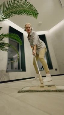 Orta yaşlı bir kadın, büyük pencereleri ve saksısı olan modern bir odada yerleri enerjik bir şekilde paspaslarken kulaklıkla müzik dinlemekten zevk alıyor. Yüksek kalite 4k görüntü