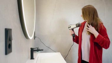 Kırmızı bornozlu genç bir kadın aynada kendine bakarken uzun saçlarını saç kurutma makinesiyle kurutuyor. Yüksek kalite 4k görüntü