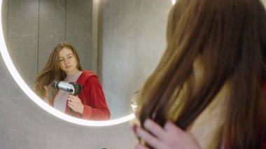 Kırmızı bornozlu genç bir kadın aynada kendine bakarken uzun saçlarını saç kurutma makinesiyle kurutuyor. Yüksek kalite 4k görüntü