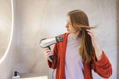 Kırmızı bornozlu genç bir kadın uzun saçlarını modern bir banyoda saç kurutma makinesiyle kurutuyor. Yüksek kalite fotoğraf