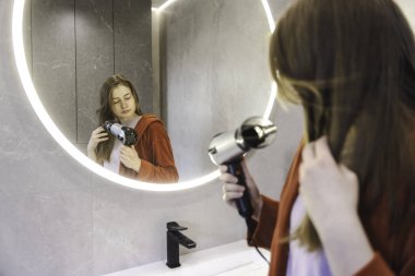 Kırmızı bornozlu genç bir kadın banyodaki aynada kendine bakarken saçını kurutma makinesiyle kurutuyor. Yüksek kalite fotoğraf