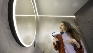 Kırmızı bornozlu genç bir kadın modern bir banyoda parlak yuvarlak bir aynayla uzun saçlarını saç kurutma makinesiyle kurutuyor. Yüksek kalite fotoğraf