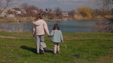 Kırsal kesimde güneşli bir bahar gününde el ele tutuşan ve sakin bir göle doğru yürüyen iki genç kız. Yüksek kalite 4k görüntü