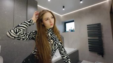 Zebra giysili genç bir kadın modern banyo içinde düşünceli bir şekilde ayakta dururken uzun saçlarına dokunuyor. Yüksek kalite 4k görüntü