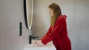 Kırmızı bornozlu genç bir kız modern bir banyo lavabosunda yüzünü suyla yıkıyor. Aydınlatılmış bir aynayla. Yüksek kalite 4k görüntü