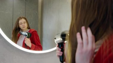 Kırmızı bornozlu genç bir kadın, yuvarlak aynadaki yansımasına bakarken saçını saç kurutma makinesiyle kurutuyor. Yüksek kalite 4k görüntü