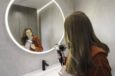 Kırmızı bornozlu genç bir kadın saç kurutma makinesi kullanıyor ve parlak parlak bir banyoda yuvarlak aynanın önünde saçını şekillendiriyor. Yüksek kalite fotoğraf