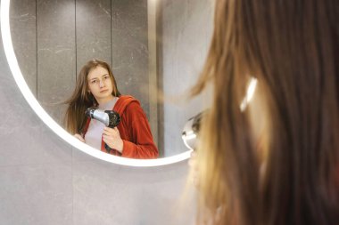 Saçını kurutma makinesiyle kurutan kırmızı bornozlu genç bir kadın modern bir banyoda parlak bir aynaya bakıyor. Yüksek kalite fotoğraf