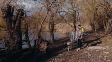 Yaşlı bir kadın, yetişkin kızı ve torunu güneşli bir bahar gününde Golden Retriever 'larıyla nehir kenarında manzaralı bir yolda yürüyorlar. Aile huzurlu doğa ortamının tadını çıkarıyor. Yüksek kalite