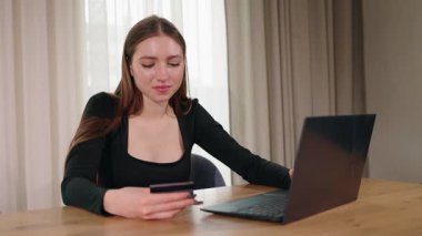 Odaklanmış genç bir kadın, kapalı bir yerdeki ahşap bir masadan online alışveriş yaparken bir dizüstü bilgisayardan kredi kartı bilgilerine giriyor. Yüksek kalite 4k görüntü