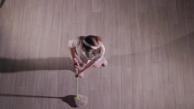 Temizlik yaparken paspasla dans eden genç bir kadın, yukarıdan yakalanmış. Beyaz bir tişört, pembe eşofman altı ve beyaz çoraplar giyiyor. Kulaklıkla enerjik bir şekilde hareket ediyor. Ev işlerini eğlenceli ve eğlenceli hale getiriyor.
