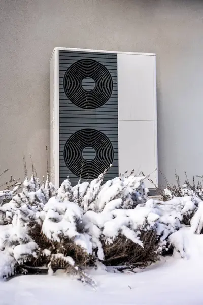 Karla kaplı çalılarla çevrili gri bir duvara monte edilmiş, kışın enerji verimliliğini ön plana çıkaran modern bir açık hava pompasının yakın çekimi. Yüksek kalite fotoğraf