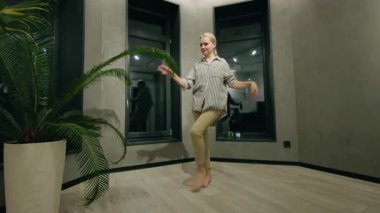 Orta yaşlı bir kadın modern bir odada çıplak ayakla dans ediyor. Büyük pencereleri ve saksısı olan bir kadın. Kaygısız ve neşeli bir anın tadını çıkarıyor. Yüksek kalite 4k görüntü