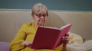 Gözlüklü yaşlı kadın yatakta kitap okuyor, hardal sarısı bir elbise giyiyor, düşünceli bir ifadeyle. Yüksek kalite 4k görüntü