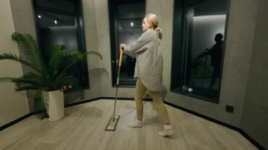 Orta yaşlı bir kadın evde paspas ile zarif bir şekilde dans ediyor. Temizliği modern bir odada eğlenceli bir ana çeviriyor. Yüksek kalite 4k görüntü
