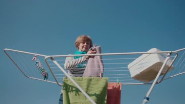 Orta yaşlı bir kadın arka bahçedeki kurutma askısına çamaşır asıyor. O çizgili bir gömlek ve mavi bir eşarp giyiyor, gri bir havlu kaldırıyor. Çamaşır sepetinin yanında, güneşte kurumuş renkli havlular var.