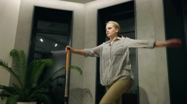 Genç bir kadın, modern minimalist bir iç mekanda elinde paspasla eğlenceli bir şekilde dans ediyor. Yüksek kalite 4k görüntü