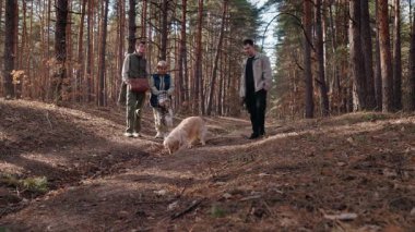 Yaşlı bir kadın ve iki yetişkin torunu gülümseyerek izlerken bir Golden Retriever kumda bir çukur kazıyor. Çam ormanında durup, köpeklerin eğlencesinin ve yaramazlığının tadını çıkarıyorlar.