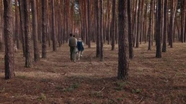 Yaşlı bir kadın iki yetişkin torunu ve bir köpekle çam ormanında yürüyor. Uzun ağaçlar ve toprak örtüsüyle çevrili doğal bir yol boyunca yürürler, huzurlu bir doğa yürüyüşünün tadını çıkarırlar..