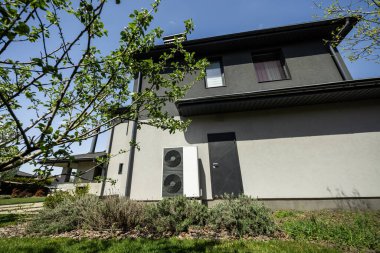 Çift pervaneli ısı pompası konut duvarına bağlı, taze yeşillik ve peyzaj ile çevrili, akıllı enerji kullanımını sembolize eden ve eko-bilinçli ev tasarımı