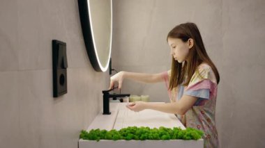 Genç kız ellerini yıkamaya hazırlanırken modern bir banyoda musluğu açıyor. Yüksek kalite 4k görüntü