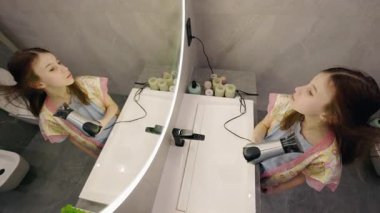 Genç bir kız, saçını büyük bir aynanın önünde saç kurutma makinesiyle kurutuyor, modern bir banyoda yansımasını en üst görüntüden gösteriyor. Yüksek kalite 4k görüntü