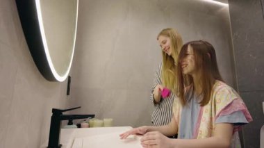 Genç kız gülümserken ablası modern bir banyoda saçını tarıyor, neşeli bir sabah rutini. Yüksek kalite 4k görüntü