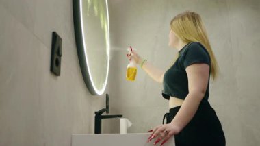 Genç bir kadın modern bir banyoda yuvarlak bir aynayı temizliyor sprey boya şişesiyle, ev işleriyle meşgul. Yüksek kalite 4k görüntü