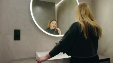 Sarışın bir kadın lavaboda duruyor ve parlak modern bir banyoda dişlerini fırçalıyor. Yüksek kalite 4k görüntü