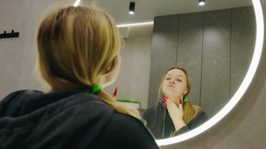Genç sarışın kadın modern bir banyoda yıkandıktan sonra temiz yüzünü aynada inceliyor. Yüksek kalite 4k görüntü