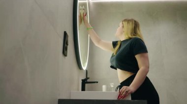 Sarışın bir kadın parlak fayanslı bir banyoda yanan yuvarlak bir aynayı temizliyor. Yüksek kalite 4k görüntü