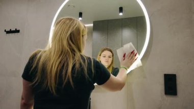 Genç sarışın bir kadın modern bir banyoda duvarları fayanslarla aydınlatılmış yuvarlak bir aynayı siliyor. Yüksek kalite 4k görüntü