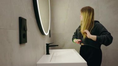 Genç sarışın bir kadın banyoda, parlak bir aynanın önünde saçına dokunuyor ve onu inceliyor. Yüksek kalite 4k görüntü