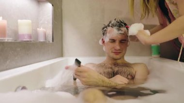 Bir kadın mum ışığında saçına şampuan dökerken köpüklü bir küvette rahatlayan gülümseyen genç bir adam. Yüksek kalite 4k görüntü