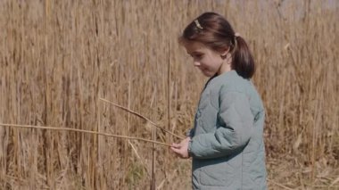 Mavi ceketli, elinde kuru kamış tutarak gülümseyen küçük kız kırsal alanda güneşlenirken. Yüksek kalite 4k görüntü