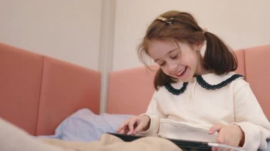 Küçük kız yatakta tablet kullanırken neşeyle gülüyor, rahat bir odada eğlenceli ve neşeli bir dijital anın tadını çıkarıyor. Yüksek kalite 4k görüntü