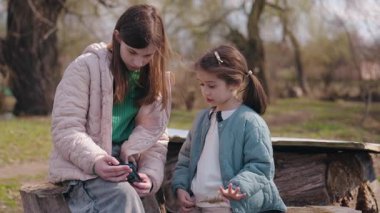 Sıcak ceketli iki kız kardeş, açık havada ağaç kütüklerine oturup güneşli bir bahar gününde birlikte telefona bakıyorlar. Yüksek kalite 4k görüntü