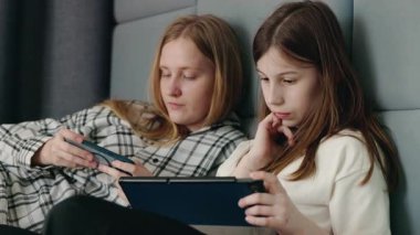 İki genç kız yan yana uzanmış, tamamen akıllı telefonlarına ve tablet ekranlarına dalmış, kapalı bir ortamda. Yüksek kalite 4k görüntü