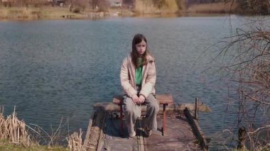 Tahta bir iskelede tek başına oturan genç bir kız, sakin bir bahar gününde düşünceli bir şekilde göle bakıyor. Yüksek kalite 4k görüntü