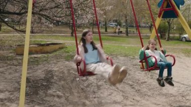 Genç bir kız ve küçük bir çocuk parkta salıncakta oynuyorlar, ılık bir bahar gününde birlikte dışarıda vakit geçiriyorlar. Yüksek kalite 4k görüntü