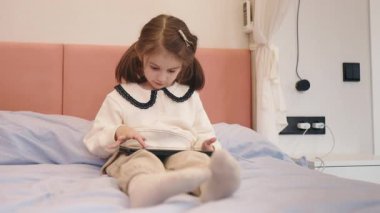 Küçük kız bir tablet kullanarak rahat bir şekilde yatakta oturuyor, rahat ve rahat bir yatak odası ortamında. Yüksek kalite 4k görüntü
